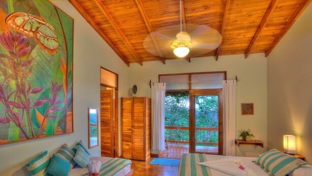 Ein Zimmer mit zwei Betten, einem Deckenventilator, einem großen Fenster und einem Bild an der Wand.