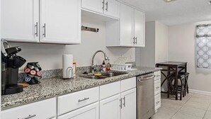 Une cuisine compacte avec des meubles blancs, un plan de travail en granit, un lave-vaisselle et un petit coin repas comprenant une table et des chaises.