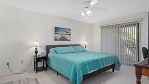 Une chambre à coucher avec un lit, deux tables de chevet avec des lampes, un ventilateur de plafond, une fenêtre avec des stores et un tableau au mur.