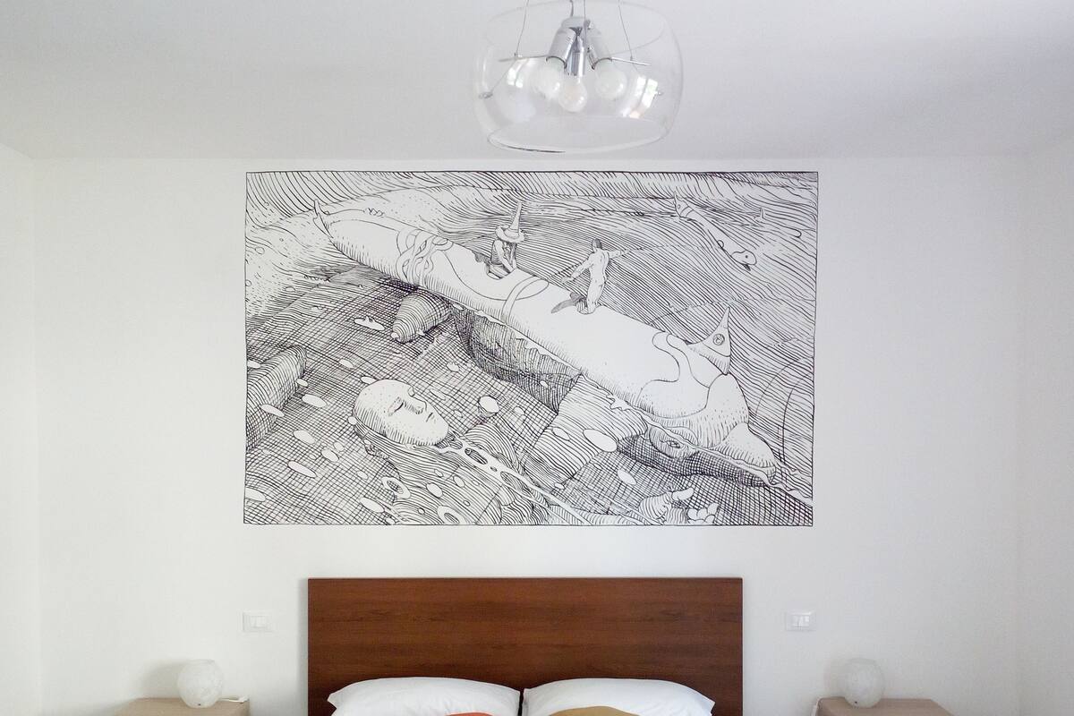 A bedroom with a wooden headboard, a bed with white and brown pillows, a wall-mounted abstract art piece, two bedside tables with lamps, and a hanging light fixture.