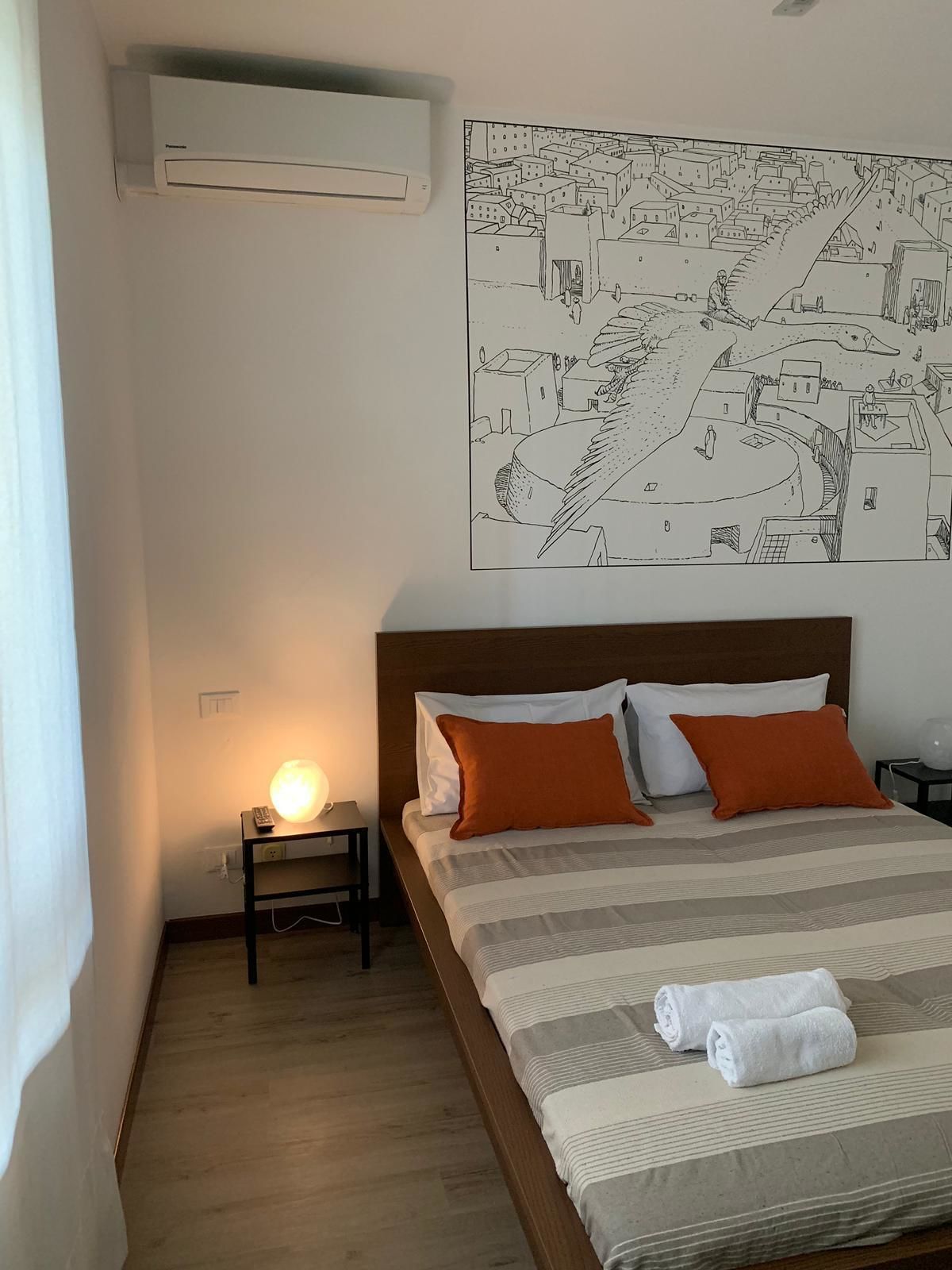 A hotel room with a bed, a nightstand with a lamp, a wall-mounted air conditioner, and a cityscape artwork.