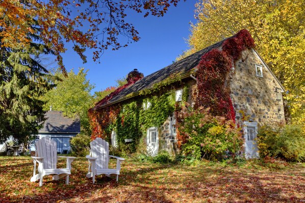 Un bâtiment en pierre recouvert de lierre et orné de feuilles d’automne, deux chaises Adirondack blanches et un ciel bleu clair.