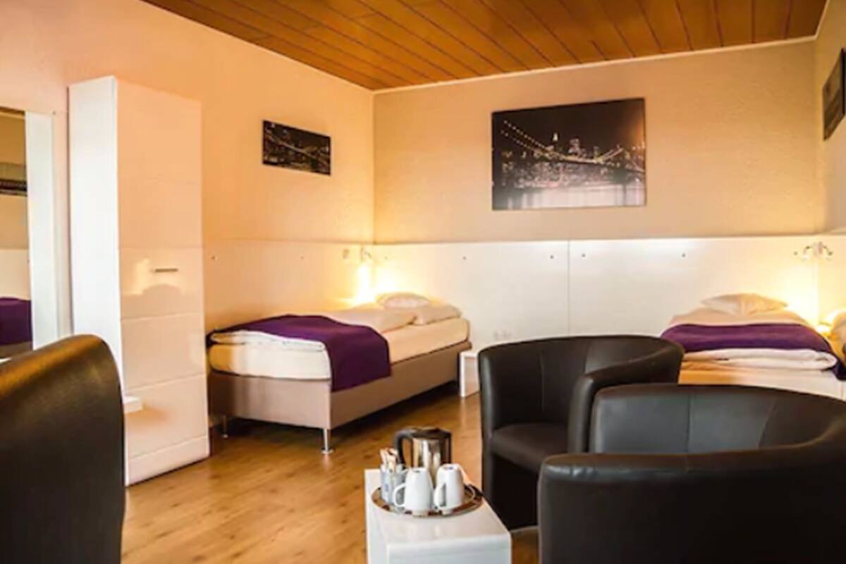 Une chambre d’hôtel avec deux lits, un canapé, une table basse avec des tasses et une bouilloire, ainsi que deux lampes fixées au mur.