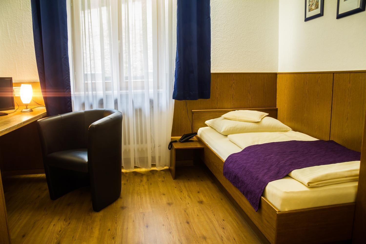 Ein Hotelzimmer mit einem Holzbett, einem Schreibtisch mit Lampe, einem Sessel und einem Fenster mit durchscheinenden Vorhängen.