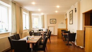 Um espaço de refeições de hotel com várias mesas e cadeiras, janelas amplas com cortinas, um balcão de recepção e um armário de madeira.