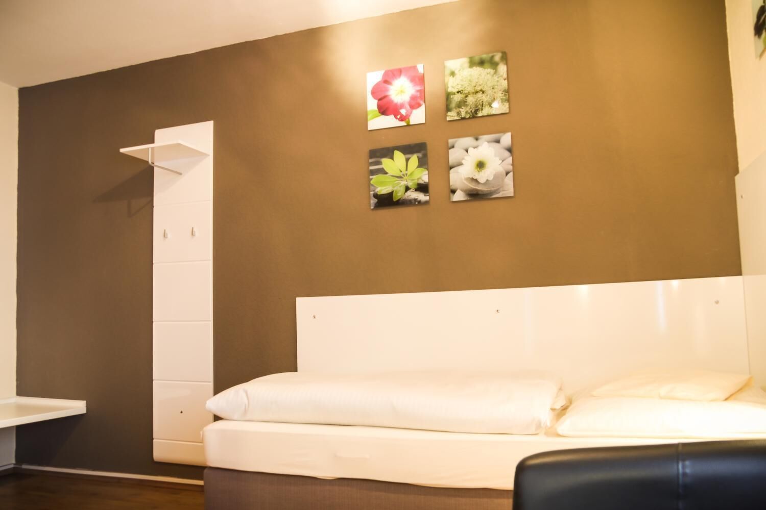 Quarto com cama branca, mesa de cabeceira branca, parede marrom e quatro quadros emoldurados.