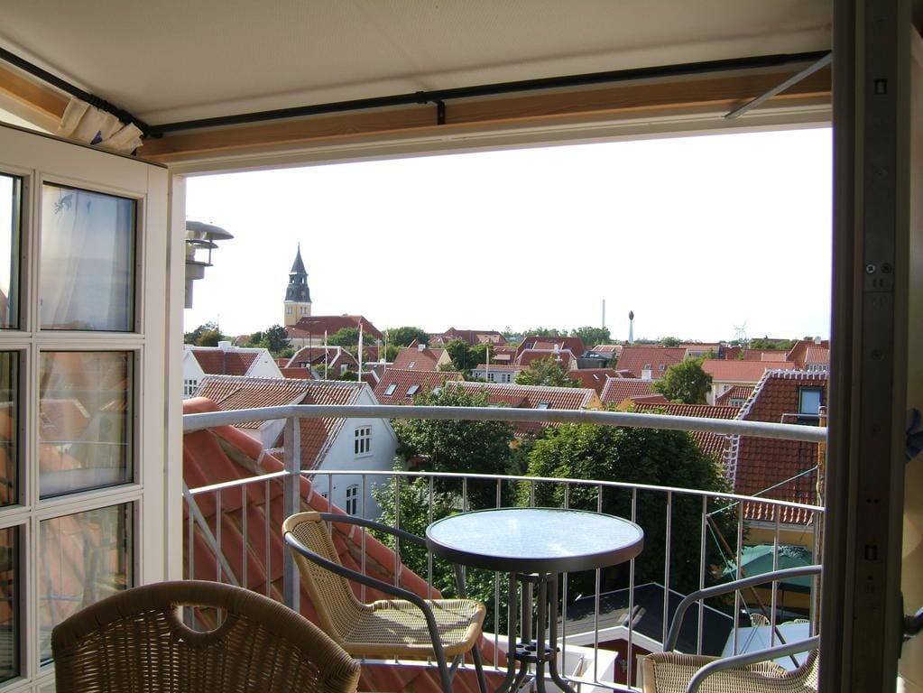 A balcony with wicker chairs and a small table, overlooking a town with red-roofed buildings and a church steeple.