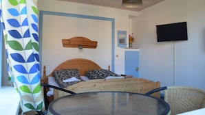Een kamer met een bed, een rond glazen tafeltje, een rieten stoel, een houten hoofdbord, een wandgemonteerde televisie en een deur met een blauwe lijst.