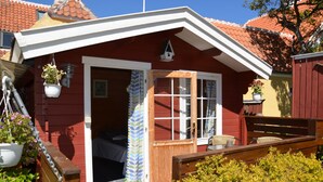 Een rode houten woning met een witte deur en ramen, een houten terras en potplanten.