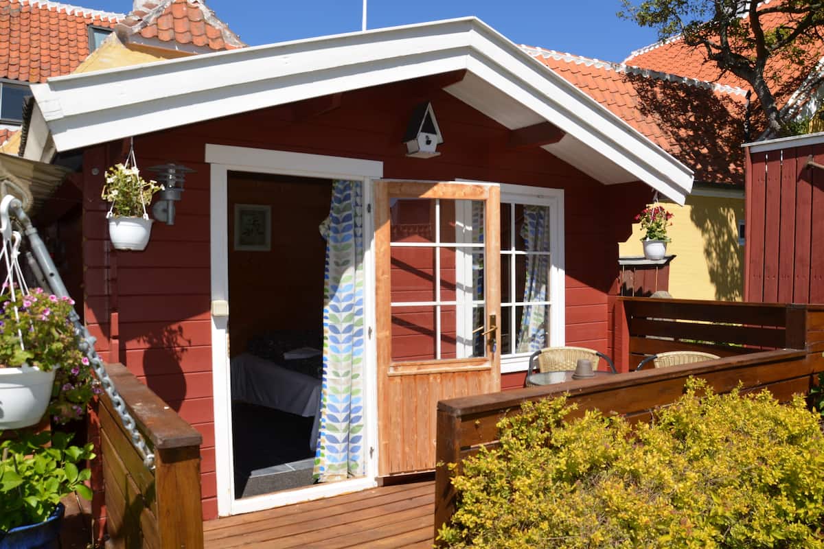 A red wooden house with a white door and windows, a wooden deck, and potted plants.