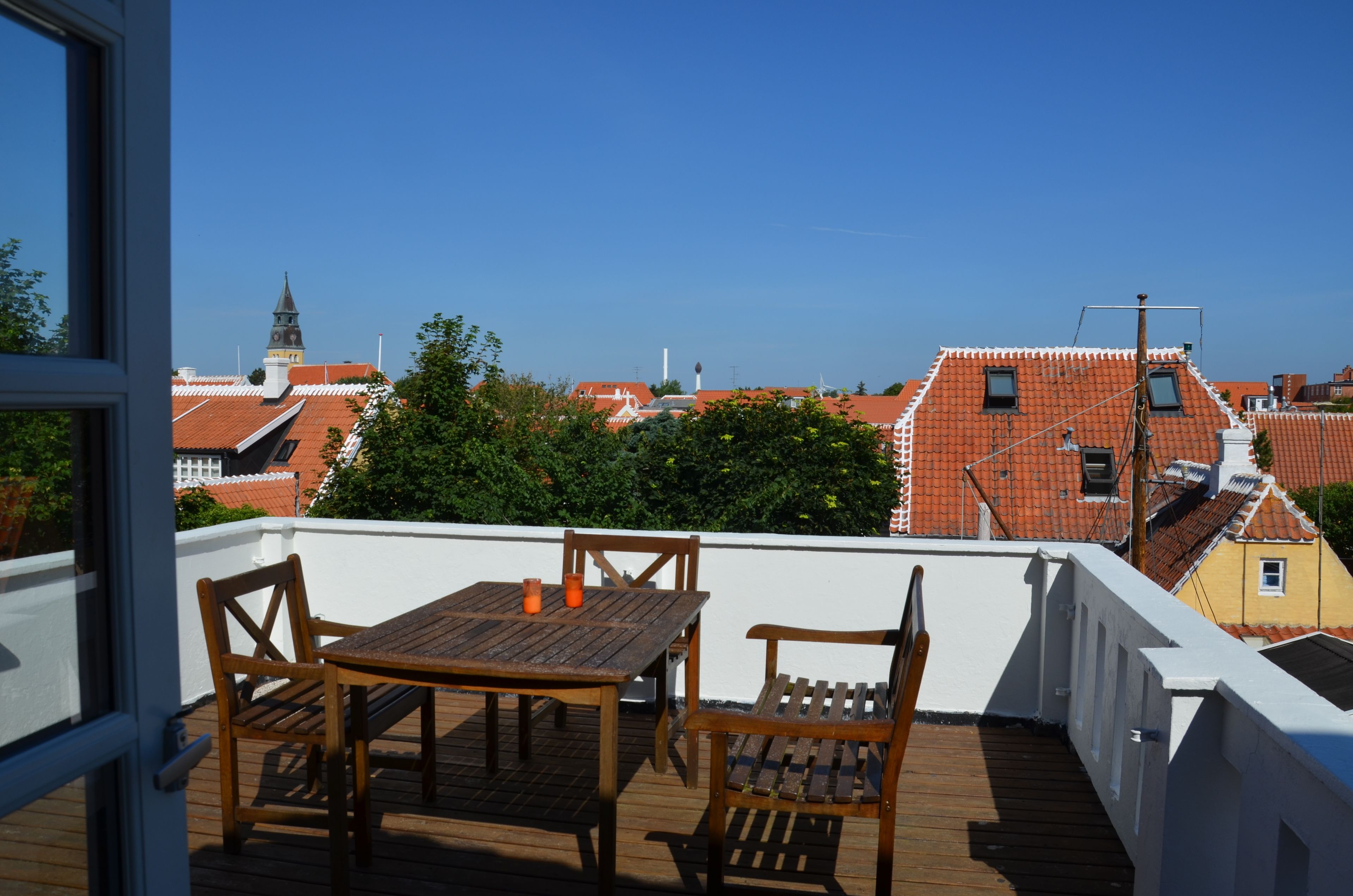 A wooden deck with a table and chairs, overlooking a cityscape with red-roofed buildings and a church steeple.