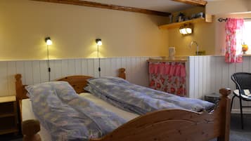Una cama de madera con ropa de cama floral, una mesita de noche, una silla, un estante con utensilios de cocina y una ventana con cortinas.