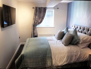 A bedroom with a bed, a TV mounted on the wall, a window with blinds, and a curtain. - The Place Blackpool (Blackpool)
