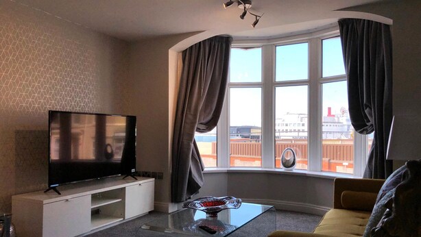 A modern living room with a flat-screen TV on a white cabinet, a glass coffee table with a decorative item, a yellow sofa, and a view of a building through large windows with curtains. - The Place Blackpool (Blackpool)