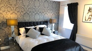 A neatly made bed with black and white bedding, two bedside tables with lamps, a patterned wallpaper, and a window with curtains. - The Place Blackpool (Blackpool)