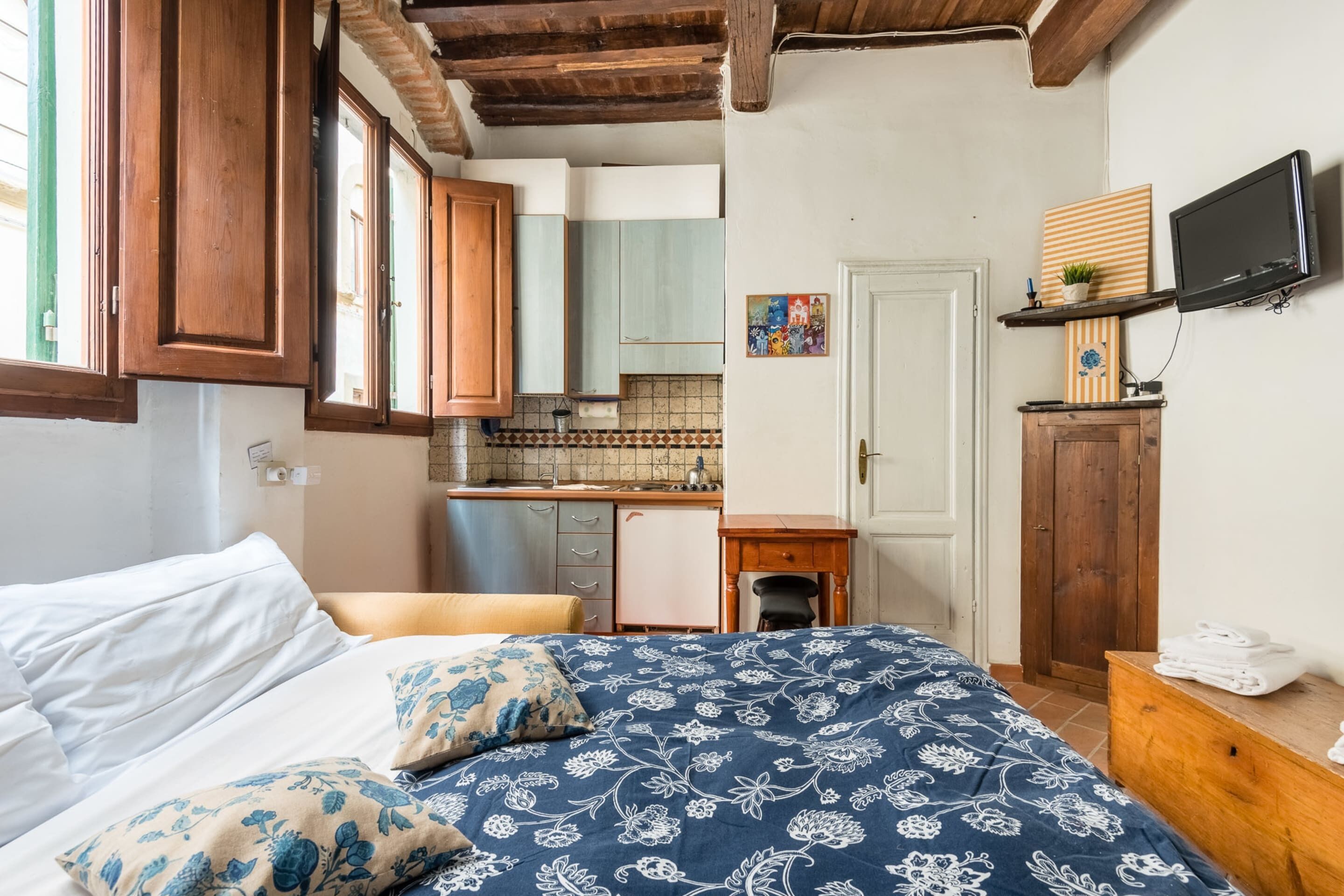 A bedroom with a bed, a wooden dresser, a television, and a kitchen area with cabinets and a tiled backsplash.