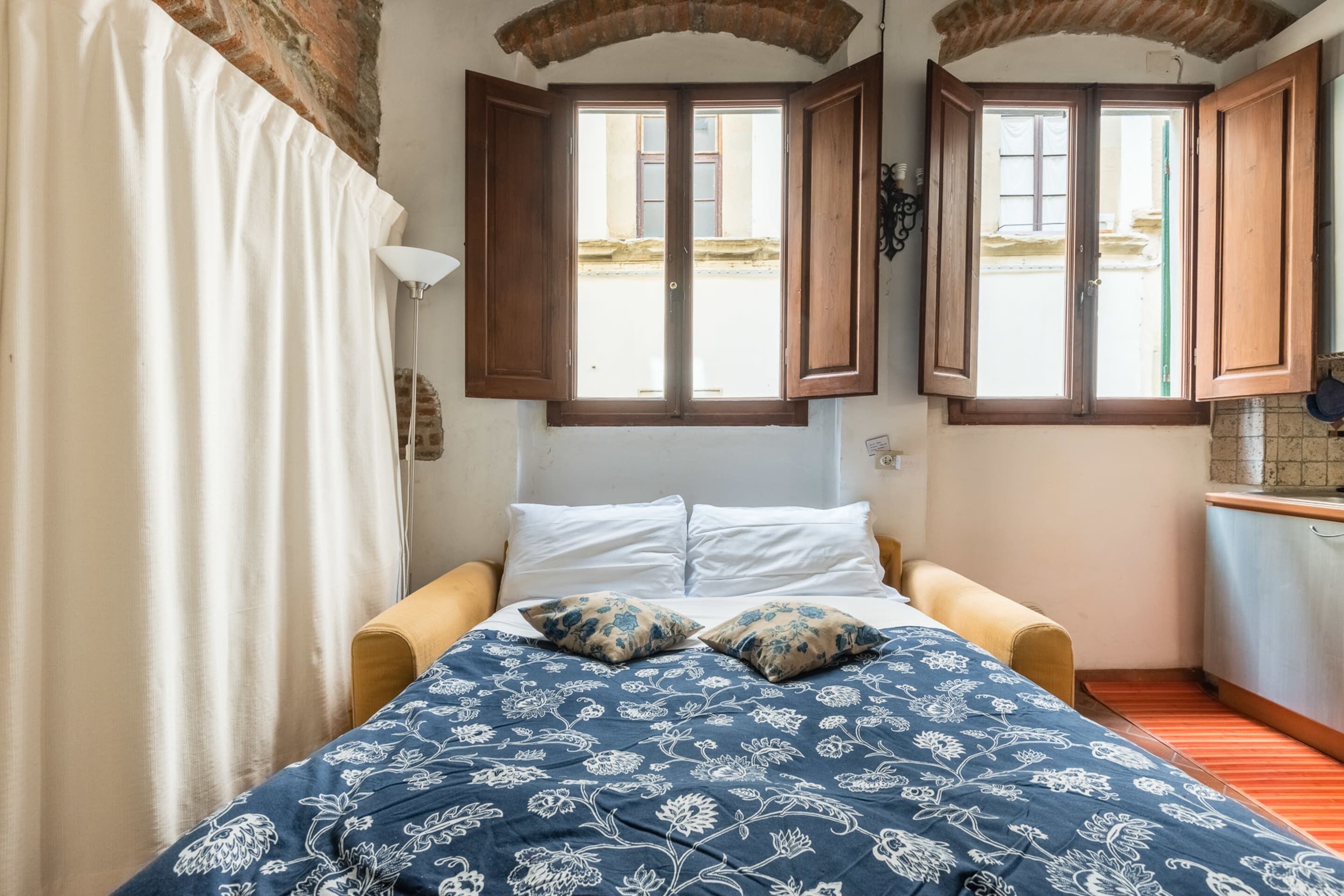 A bedroom with a bed, two pillows, a nightstand, a lamp, and a window with wooden shutters.