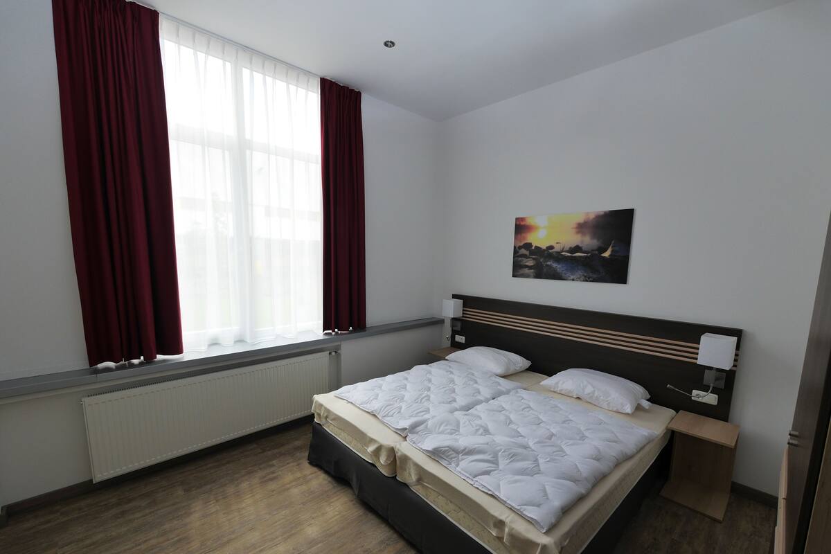 Una habitación de hotel con una cama grande, una mesita de noche, un cuadro en la pared y una ventana con cortinas.