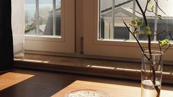 Una taza de vidrio transparente sobre un platillo, un florero de vidrio con una rama, y una ventana con vista a edificios.