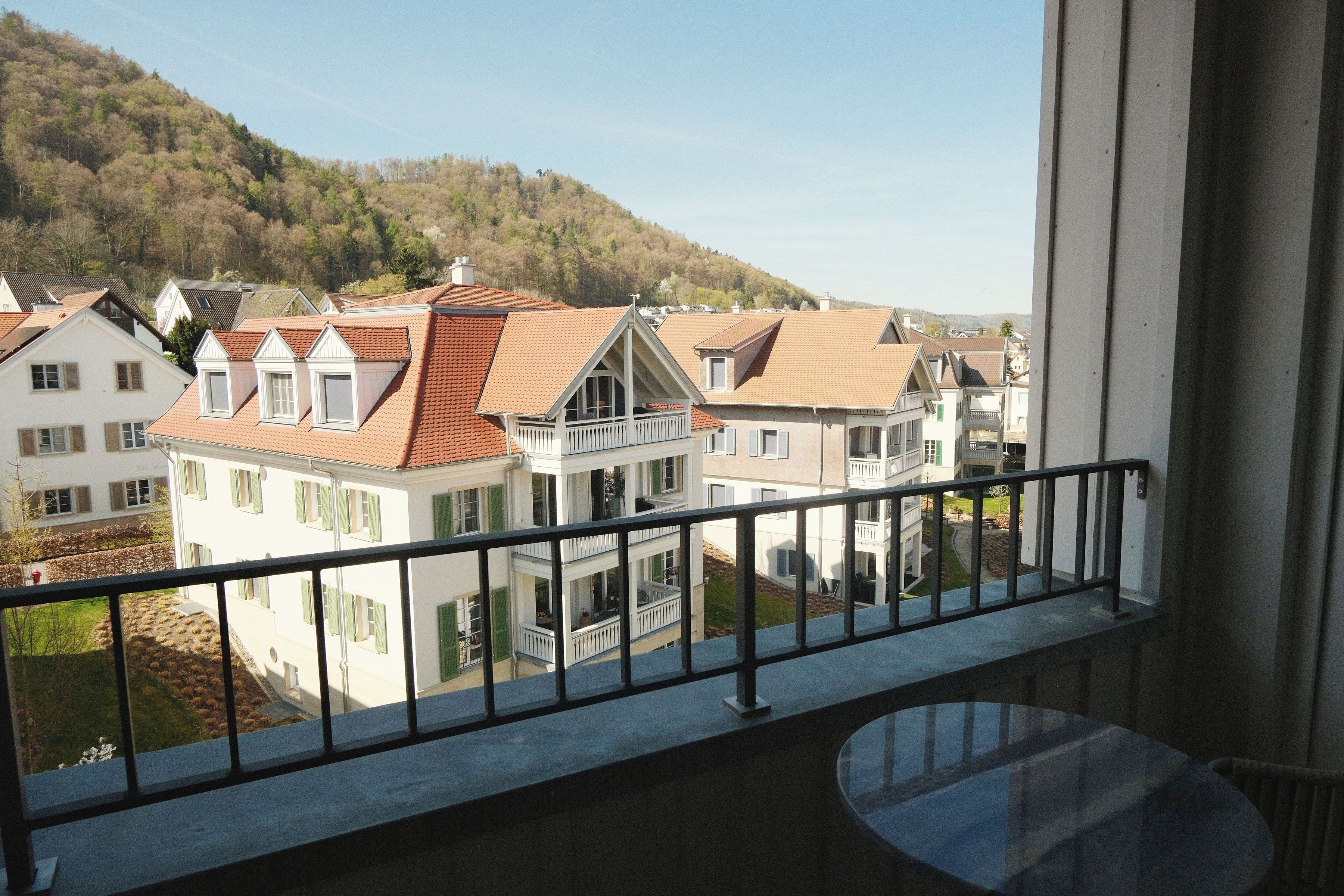 A residential building with a balcony, a view of neighboring houses, and a hillside in the background.