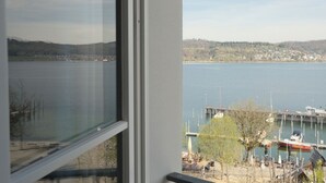 Une vue à travers une fenêtre sur un lac, avec un quai et des bateaux, une ville lointaine et un paysage vallonné.