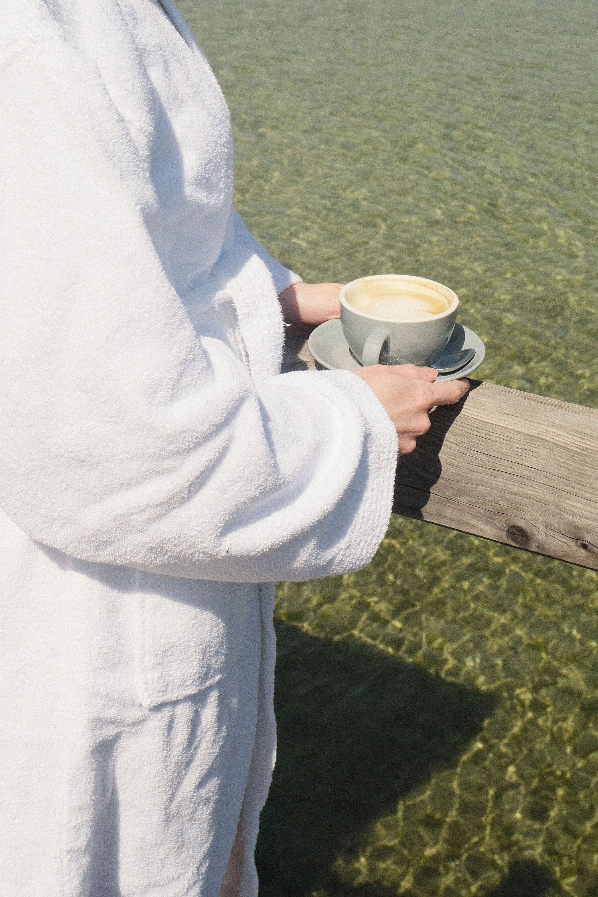 A person in a white bathrobe holding a cup of coffee on a wooden bench.