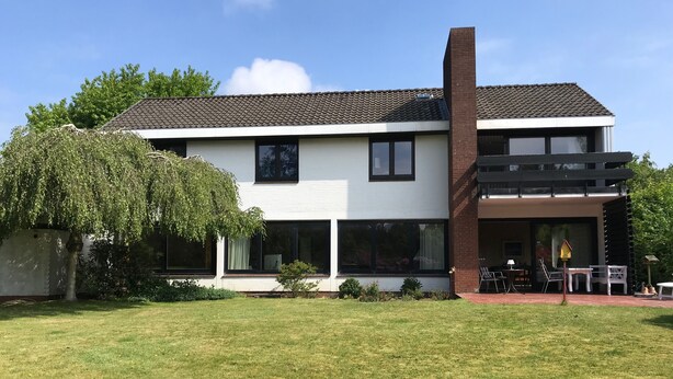 A modern two-story house with a large front lawn, a brick chimney, and a patio area with outdoor furniture. - Charming 70s house with large garden (Heide)
