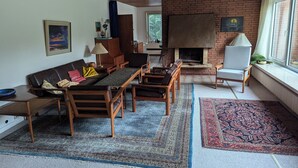 A cozy living room with a brick fireplace, wooden furniture, and a large window. - Charming 70s house with large garden (Heide)