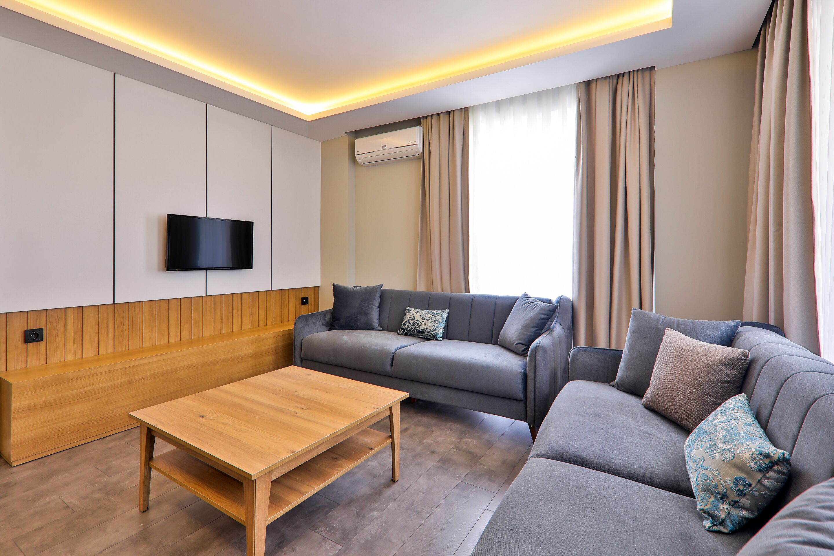 Ein modernes Wohnzimmer mit einer Couch, einem Holztisch, einem Flachbildfernseher an der Wand und einer Klimaanlage.