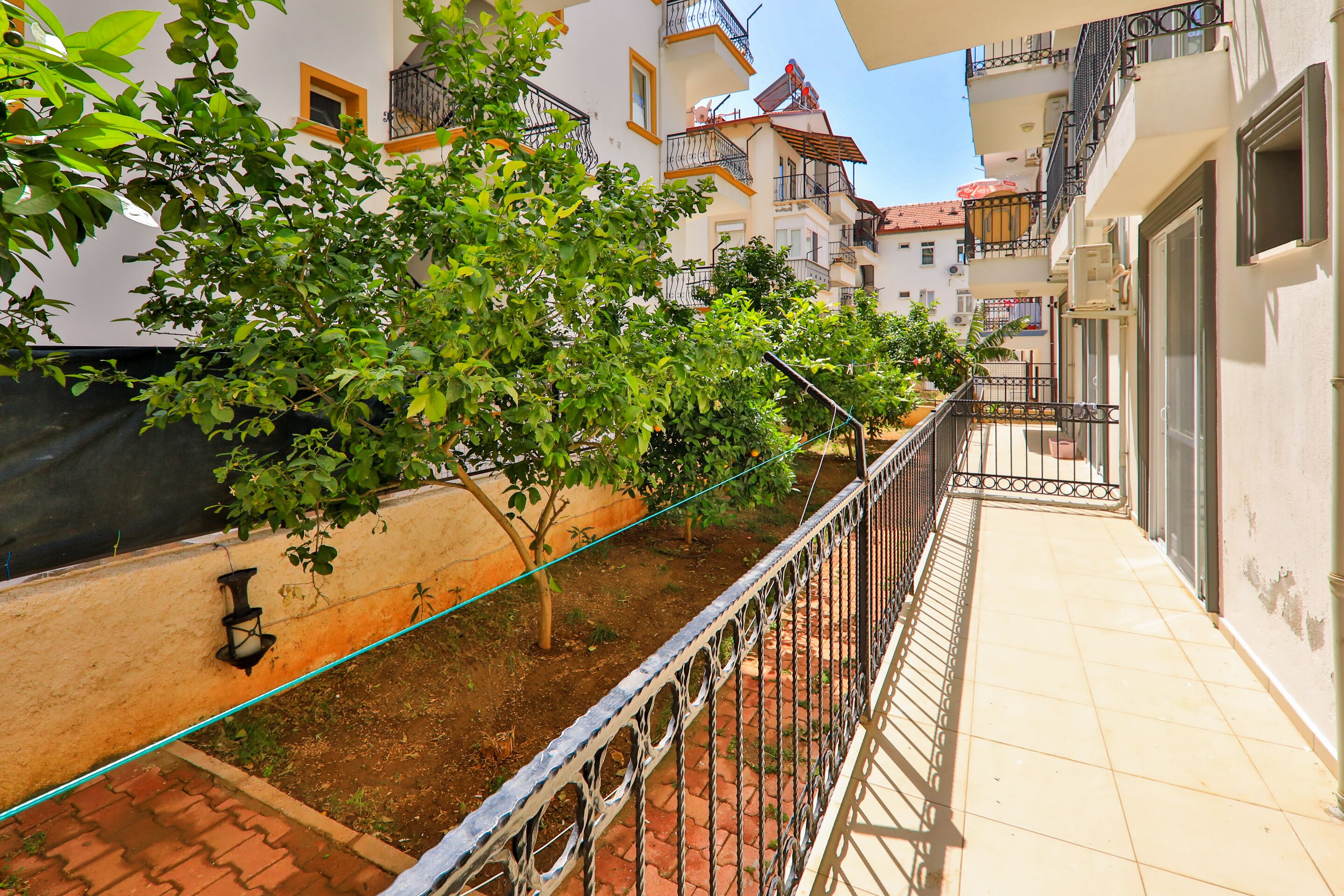 A narrow outdoor corridor with a tiled floor, metal railing, and greenery.