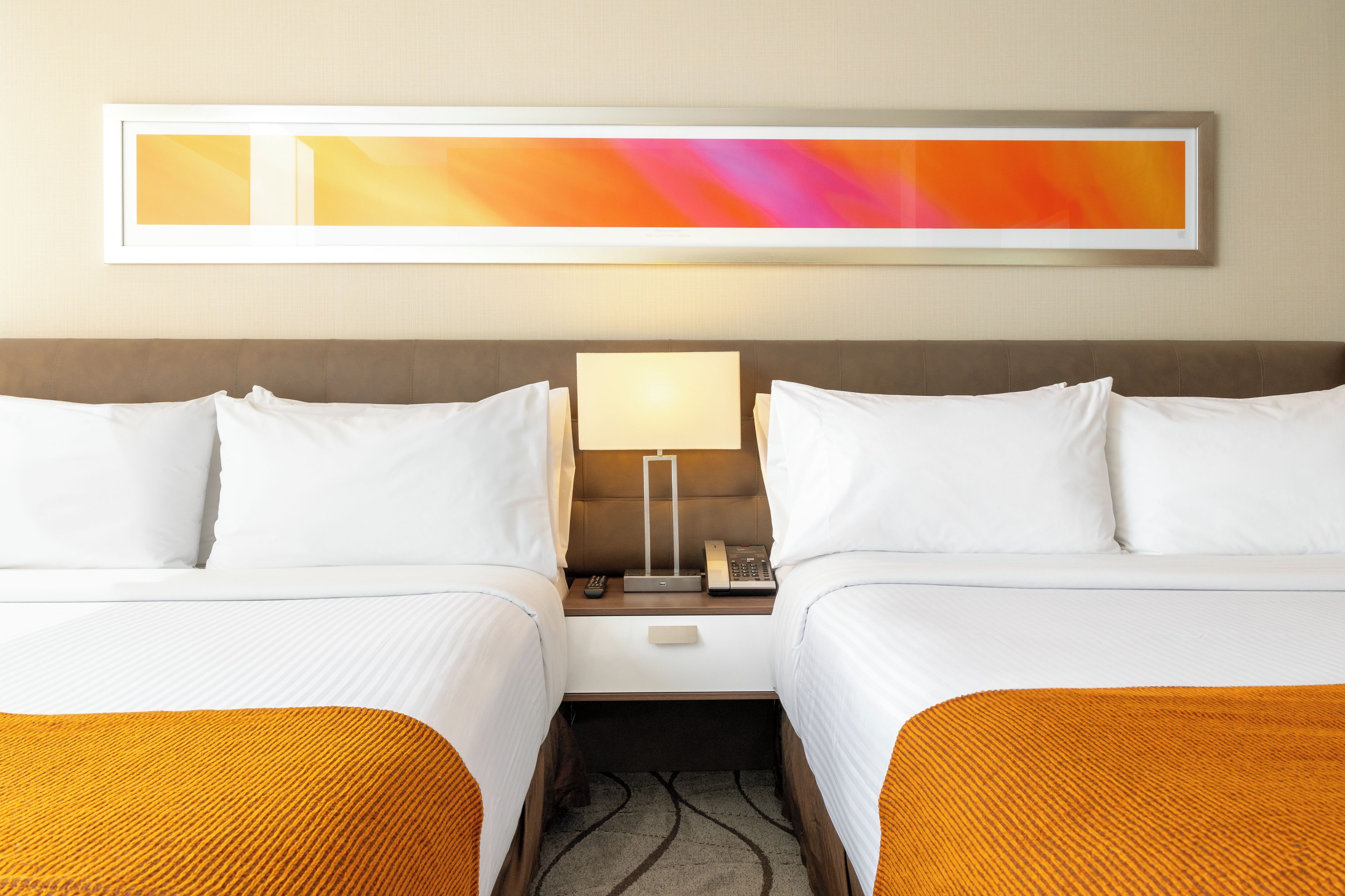 A hotel room with two beds, a nightstand with a lamp and phone, and a large abstract painting on the wall.