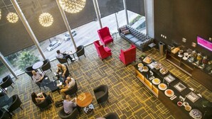 A modern hotel lobby with a geometric patterned carpet, red and black seating, a bar area with food and drink options, and large windows offering a view of the street.