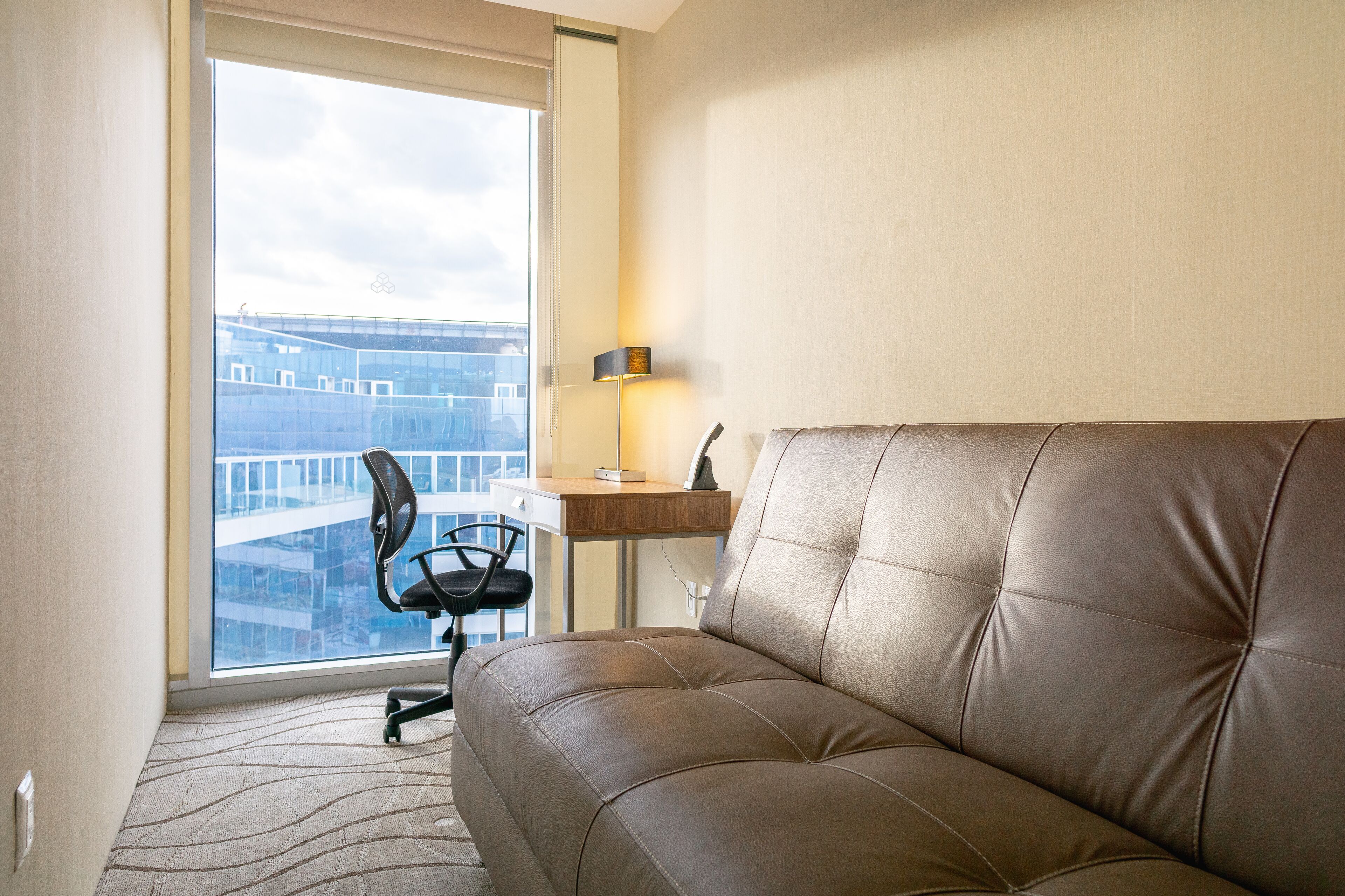 Une chambre d’hôtel moderne, dotée d’un canapé en cuir, d’un bureau et d’une chaise. Une grande fenêtre offre une vue sur le paysage urbain.
