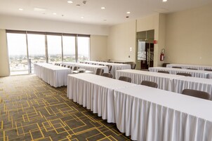Un salón de conferencias con varias mesas y sillas, ventanas amplias con vistas a la ciudad y un extintor de incendios montado en la pared.