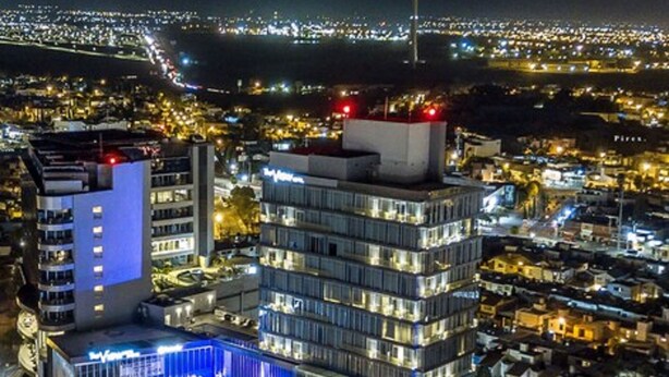 A multi-story hotel with a pool area, a rooftop terrace, and a view of the cityscape at night.