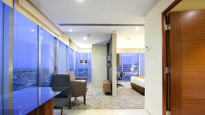A modern hotel room with a large window, a desk, a chair, a bed, and a wooden door.