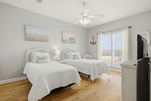 A bedroom with two beds, a ceiling fan, artwork on the walls, and a window with curtains.