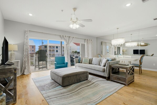 A spacious living room with a large window, a ceiling fan, a sofa, an ottoman, and a small table.