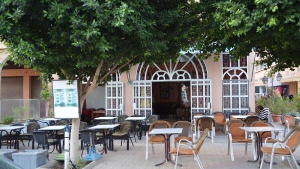 Ein Straßencafé mit Tischen und Stühlen, ein Hauseingang und ein Baum, der Schatten spendet.