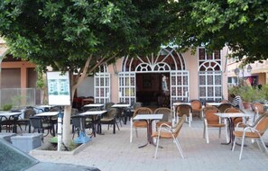 Un espace café en plein air avec des tables et des chaises, une entrée d’immeuble et un arbre qui offre de l’ombre.
