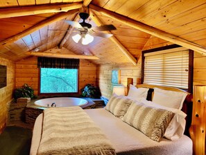 A cozy cabin bedroom with a large bed, a ceiling fan, a window with blinds, and a bathtub. - Secluded Hideaway Cabin boasting Wrap-Around Deck and Jacuzzi for two! Trail & Cave on property! (Eureka Springs)