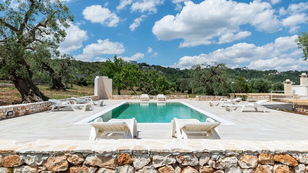 Ein rechteckiger Pool mit weißen Liegestühlen, umgeben von Steinmauern und Grünpflanzen, unter blauem Himmel.