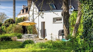 Une maison à l’extérieur blanc, avec un escalier en pierre, un coin repas comprenant une table et des chaises, et un grand arbre recouvert de lierre.