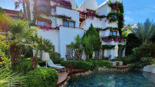 Une piscine entourée d’une végétation luxuriante et de bâtiments blancs ornés de fleurs roses.