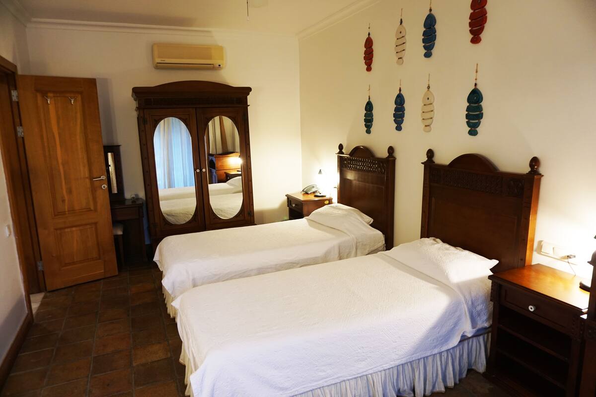 A hotel room with two beds, a wooden wardrobe, a mirror, a bedside table with a lamp, and wall decorations.