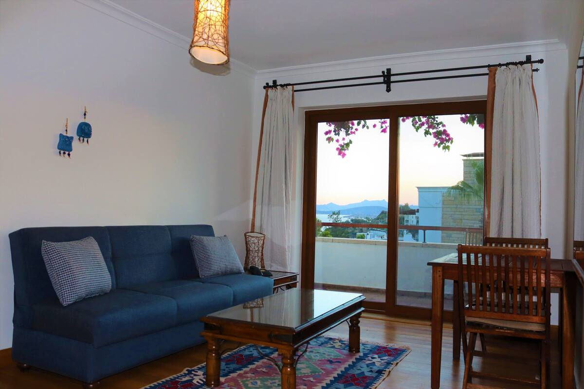A living room with a blue sofa, a glass coffee table, a wooden chair, and a view through a sliding glass door to the outside.