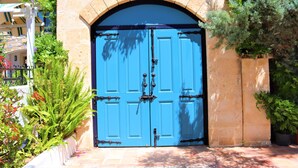 Une porte bleue avec une poignée en métal, encadrée par un arc en pierre, entourée de verdure et bordée d’un trottoir carrelé.