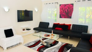 Una sala de estar moderna con una mesa de centro de vidrio, sofás negros con cojines rojos, un sillón blanco y un televisor de pantalla plana montado en la pared.