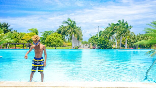 Una persona de pie en una piscina con agua azul cristalina, rodeada de palmeras y follaje.