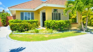 Una casa de dos pisos con techo de tejas, una puerta principal y un pequeño jardín con plantas variadas y una palmera.